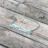 Mountain Range Bar Necklace with a silver engraved mountain silhouette on a slim bar pendant resting on a wood surface