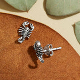 Silver scorpion shaped stud earrings on a brown suede surface with green leaves