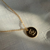 Gold chain necklace with a round pendant featuring an engraved emblem on light fabric backdrop