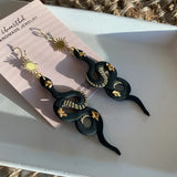 Solstice Geode Snake Earrings featuring black serpentine shapes with gold detailing and sunburst accents.