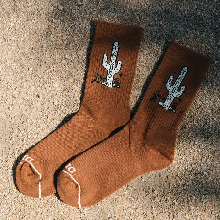 Pair of brown Cactus Taco Crew Socks with white cactus logos laid on a sandy surface.