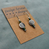 Dangling earrings with a round sunburst metal charm and green beads on a brown display card.