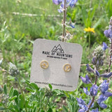 Gold mountainscape stud earrings on a beige card nestled among green plants and purple flowers.