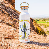 White insulated water bottle with cactus art on a rocky desert landscape.