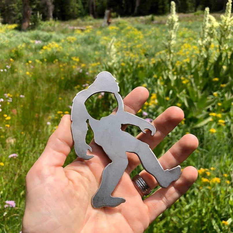 Blue metal Bigfoot Yeti bottle opener outdoors on a weathered wooden surface among wildflowers
