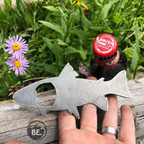 Trout shaped metal bottle opener held by fingers, with a beer bottle cap visible behind it against a garden background.