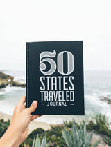 Holding a black 50 STATES TRAVELED JOURNAL with bold white text, outdoors at the coast.