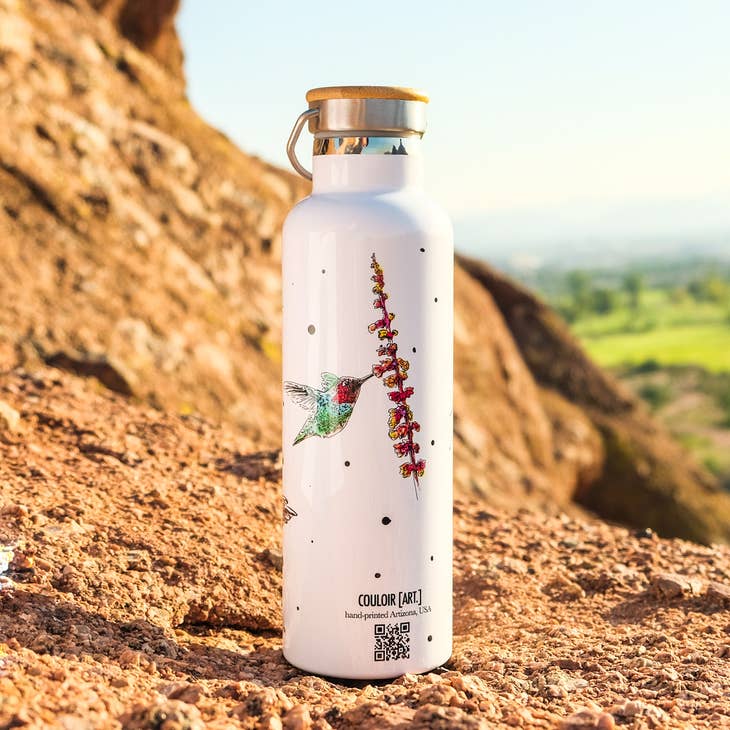 White steel water bottle with hummingbird and red flowering plant motif, wooden cap, set on a rocky hillside.