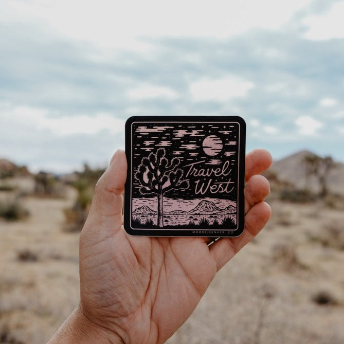 Hand holding the Travel West sticker outdoors in a desert landscape